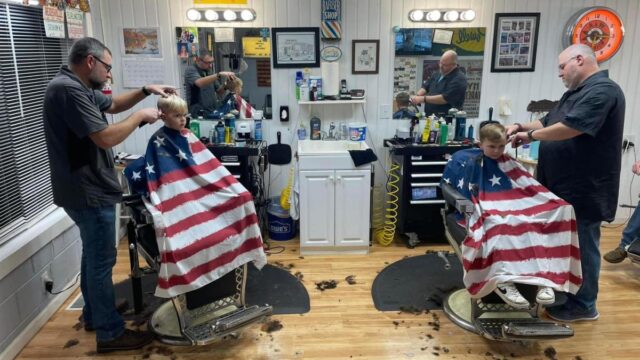 Two young boys getting haircuts side by side at a barbershop, draped in American flag smocks