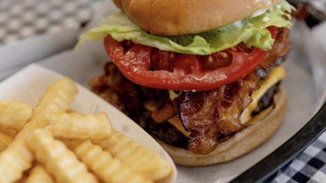 A tall bacon cheeseburger and French fries on a white plate on top of a black and white checked tablecloth