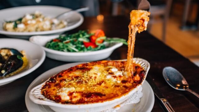 A fork lifts a cheesy bit of food from a white dish on a brown wooden table, surrounded by other dishes of food